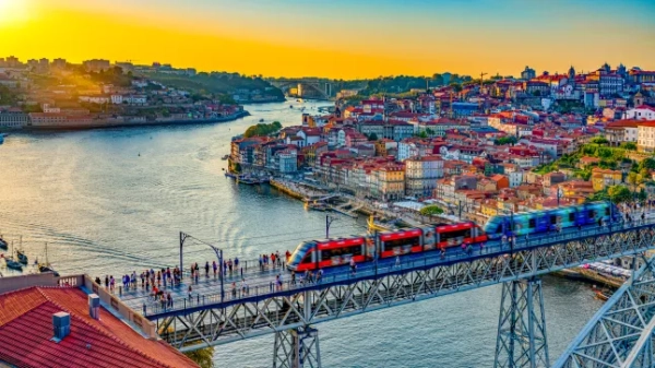 Вид у сутінках на червоно-синій поїзд, що їде мостом через річку Дору в Порту, Португалія, з будинками, що вишикувалися вздовж берега річки.
