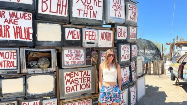 Жінка стоїть перед арт-інсталяцією з телевізорів, намальованих червоним. На ній сині шорти, біла майка та сонцезахисні окуляри.