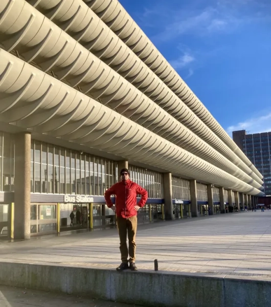 Бен Айткен у червоній куртці стоїть на стіні біля модерністської будівлі в Престоні