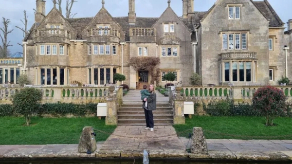 Рейчел Мосс тримає свою дитину на руках біля величного фасаду розкішного сімейного готелю Woolley Grange у Вілтширі.