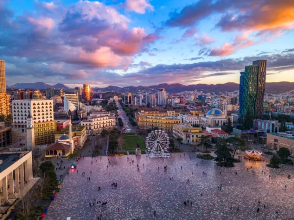 Вид на головну площу Тирани Шеші Скендербей на заході сонця, оточену будівлями та хмарочосами, на тлі гір вдалині.