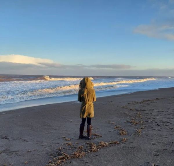 Седі Вайтлокс стоїть у пальто спиною до камери на пляжі в Лінкольнширі.