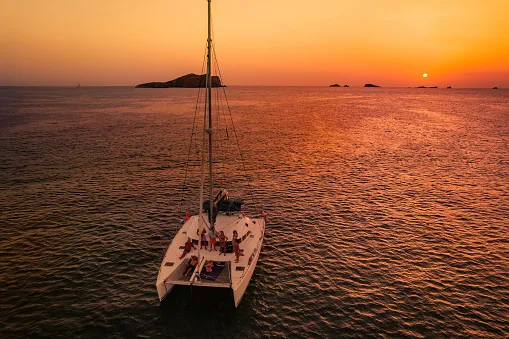 Вид з повітря на групу друзів, які відпочивають на палубі катамарана, споглядаючи захід сонця під час канікул на острові Ібіца.