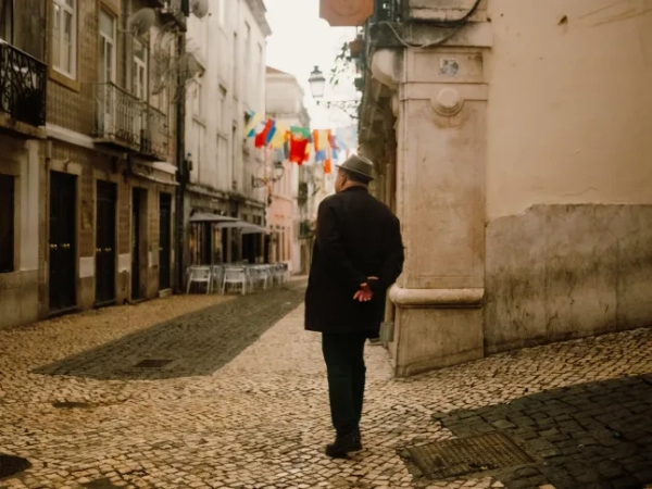 Чоловік у чорному костюмі та сірому капелюсі-котелку стоїть спиною до камери, заклавши руки за спину, на брукованій вулиці в Лісабоні.