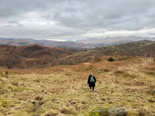Сара Хупер, зображена здалеку, йде полями та пагорбами в Озерному краї сірого хмарного дня.