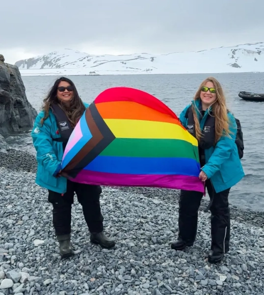 Дві жінки тримають прапор Прайду на сланцевому пляжі в Антарктиді