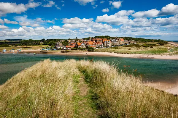 Grassy dunes in the foreground, with the incredibly quaint seaside village of Alnmouth and the mouth of the Aln River in the background, on a bright sunny day.