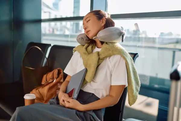 Young Asian woman sleeping in an airport terminal, wearing a neck pillow and holding a laptop, waiting for a flight