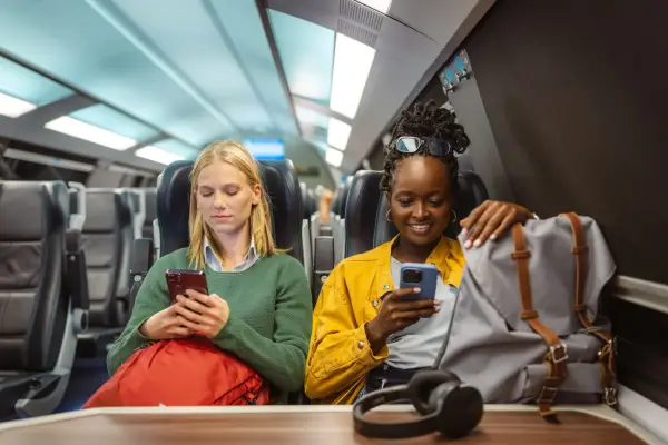 Two diverse friends ride a modern high-speed train, relaxing with backpacks and smartphones during the train ride