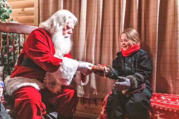 Santa Claus handing a present to a young blonde girl in a cosy room