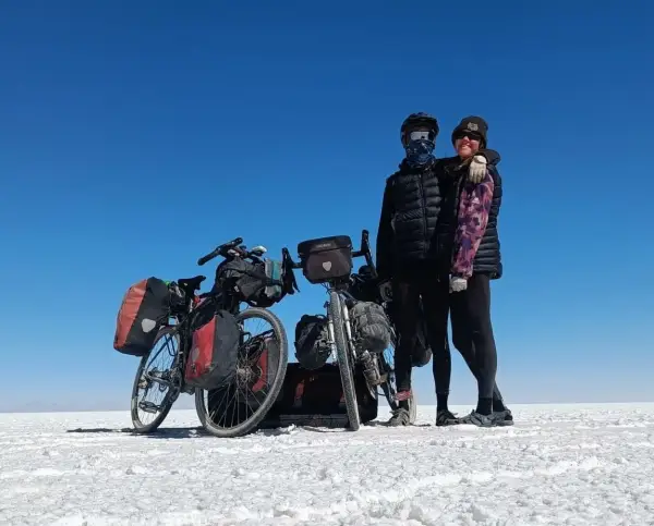 Eline and Jonny posing next to their bikes on the Salar de Uyuni salt flats in Bolivia. The ground is white, and the sky is bright blue and completely cloudless.
