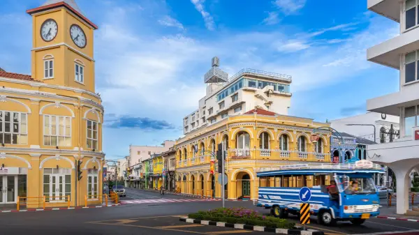 The iconic yellow clock tower and historic Chartered Bank building in Phuket Old Town, showcasing Sino-Portuguese architecture, frame a bustling intersection with a blue local bus passing by