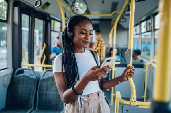 Young African american woman wearing earphones and listening to music on a smartphone while standing alone on a bus. Traveling to work and enjoying a bus ride. Portrait of a beautiful black woman.; Shutterstock ID 2189852039; purchase_order: -; job: -; client: -; other: -