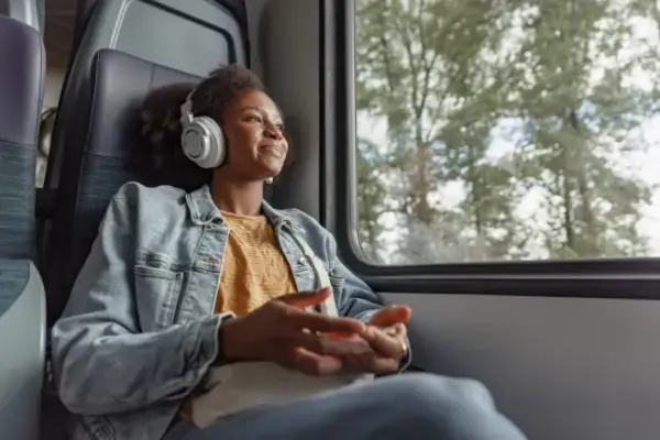 Young African American woman comfortably sitting by the train window, watching the beautiful scenery and enjoying music through her headphones.; Shutterstock ID 2567490795; purchase_order: -; job: -; client: -; other: