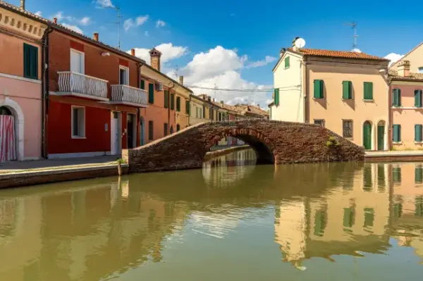 Stunning view of Comacchio featuring an ancient brick arch bridge perfectly reflecting on the calm canal. Colorful traditional houses with warm tones line the waterway under a bright summer blue sky filled with soft white clouds in daytime.