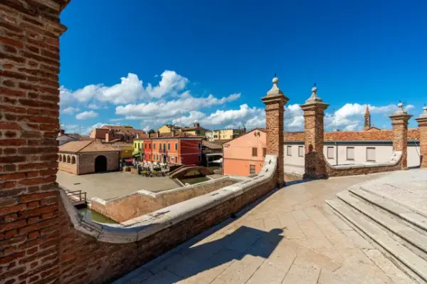 Stunning high-angle view from the Trepponti bridge overlooking the historic village of Comacchio. The shot displays red brick walls, sunny squares, traditional colorful houses, and a vibrant blue sky enriched by fluffy white clouds in daylight.