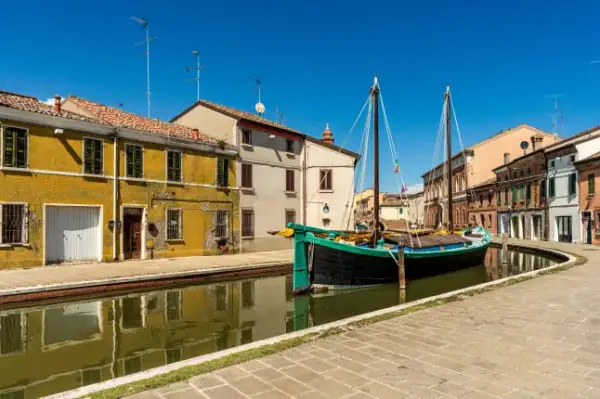 A historic wooden boat with masts moored along the main canal in the center of Comacchio. The typical traditional houses with warm yellow tones reflect on the still water surface, under a crystal-clear, deep blue summer sky without clouds.